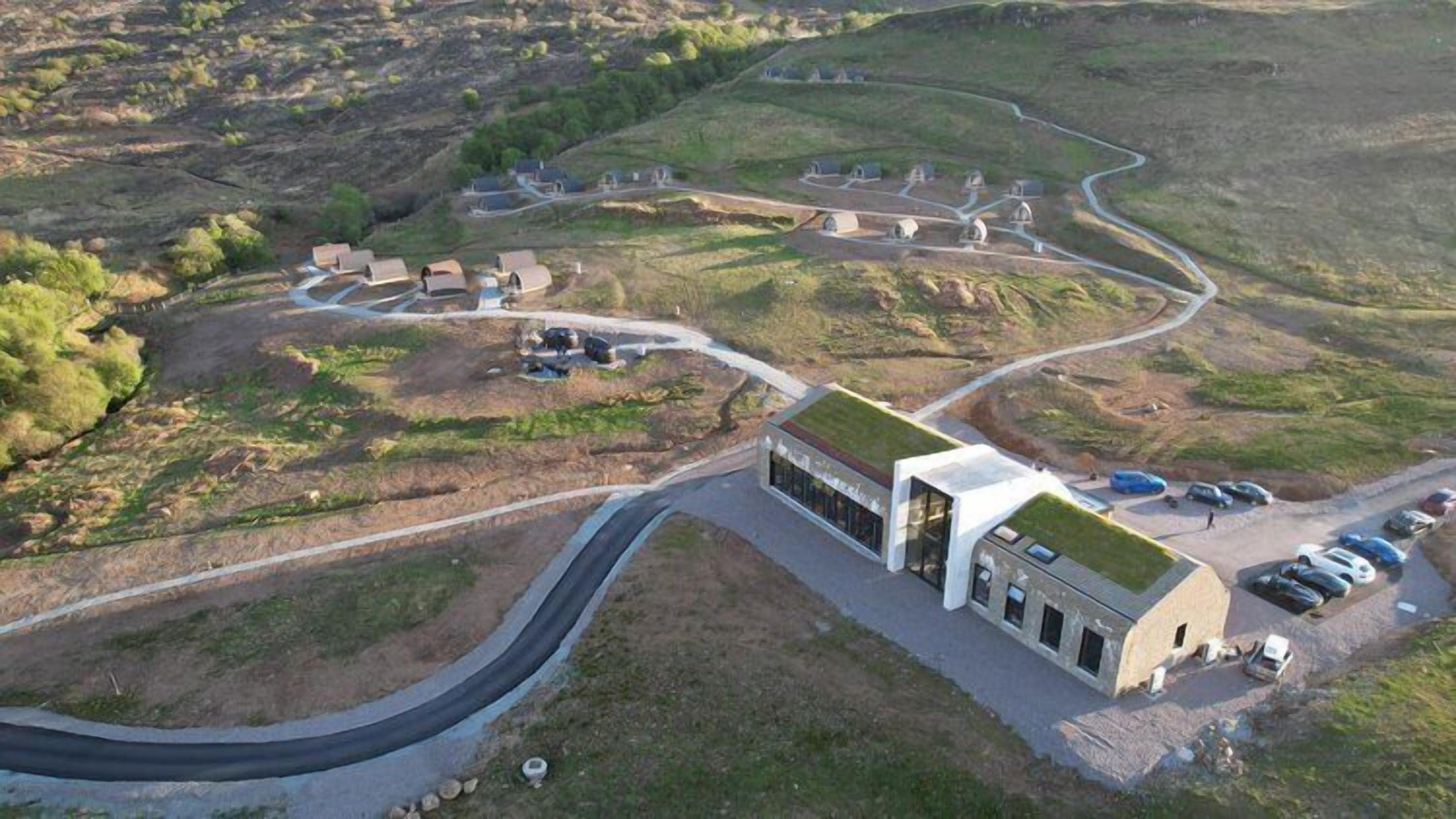 An aerial view of a modern building with a green roof, surrounded by a winding road and a series of smaller structures.