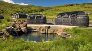 A series of wooden cabins with arched windows, a small pond with a waterfall, and a grassy hillside in the background. - Bracken Hide Hotel (Portree)