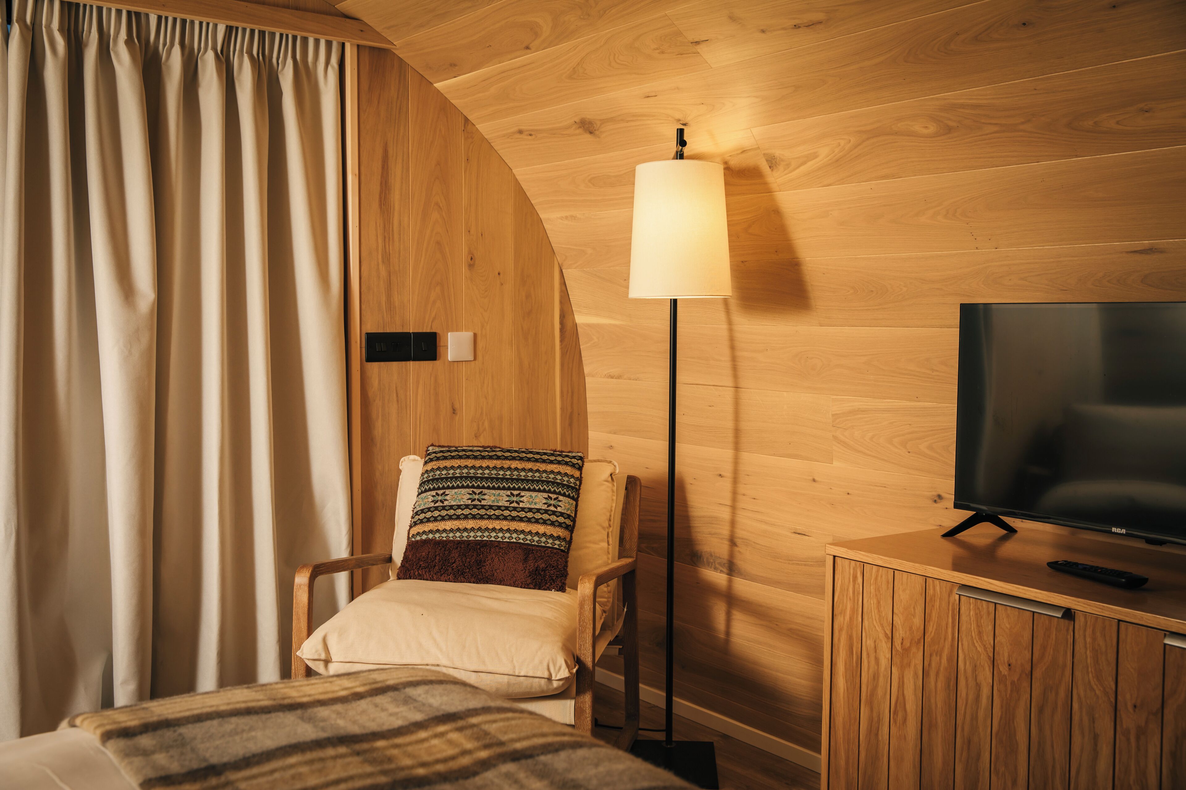 Una habitación con techo de madera, una cama con manta a rayas, una silla con cojín estampado, una lámpara de pie y un televisor sobre un mueble de madera.