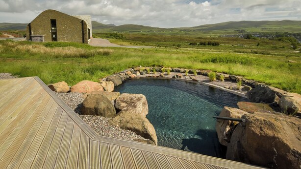 A wooden deck leading to a clear pool surrounded by rocks, with a stone building in the background. - Bracken Hide Hotel (Portree)