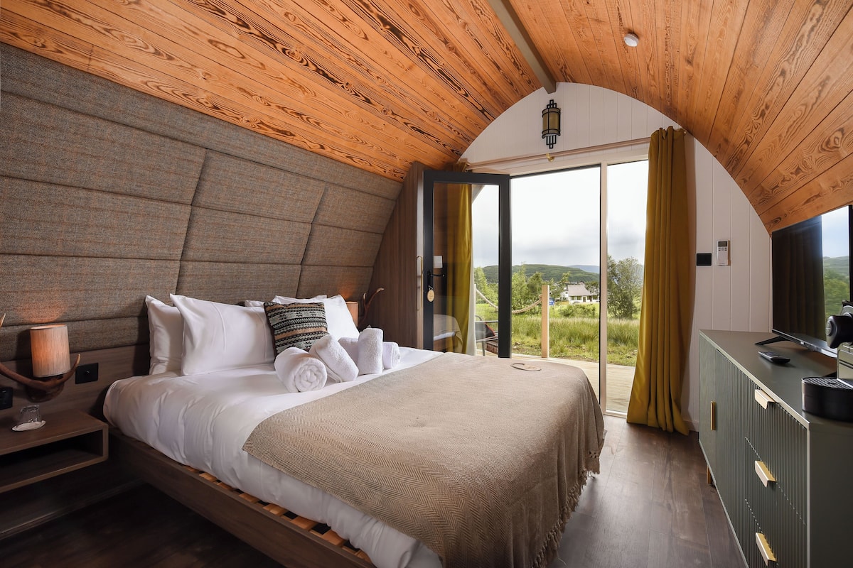 Ein Schlafzimmer mit Holzboden, einem Bett mit weißer Bettwäsche, einem Nachttisch mit Lampe und Blick ins Freie durch ein großes Fenster.