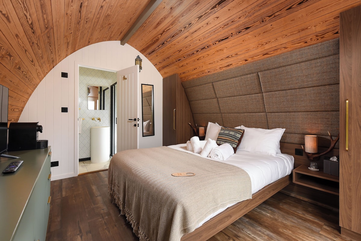 Ein Schlafzimmer mit Holzboden, einem Bett mit einer beige farbenen Decke, einem Nachttisch mit Lampe und einem Spiegel an der Wand.