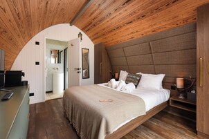 Un dormitorio con techo de madera, una cama con manta color beige, una mesita de noche con lámpara y un espejo en la pared.