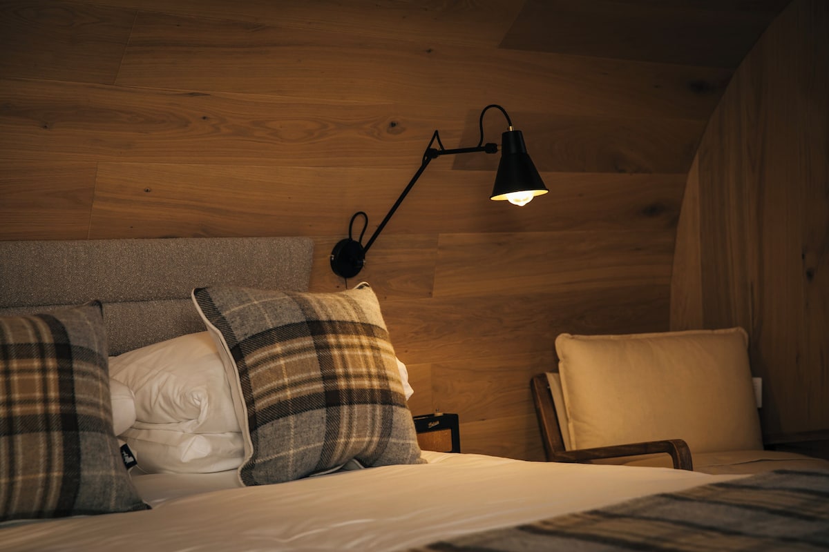 Ein gemütliches Zimmer mit Holzboden, einem Bett mit karierten und weißen Kissen sowie einem Nachttisch mit Lampe.