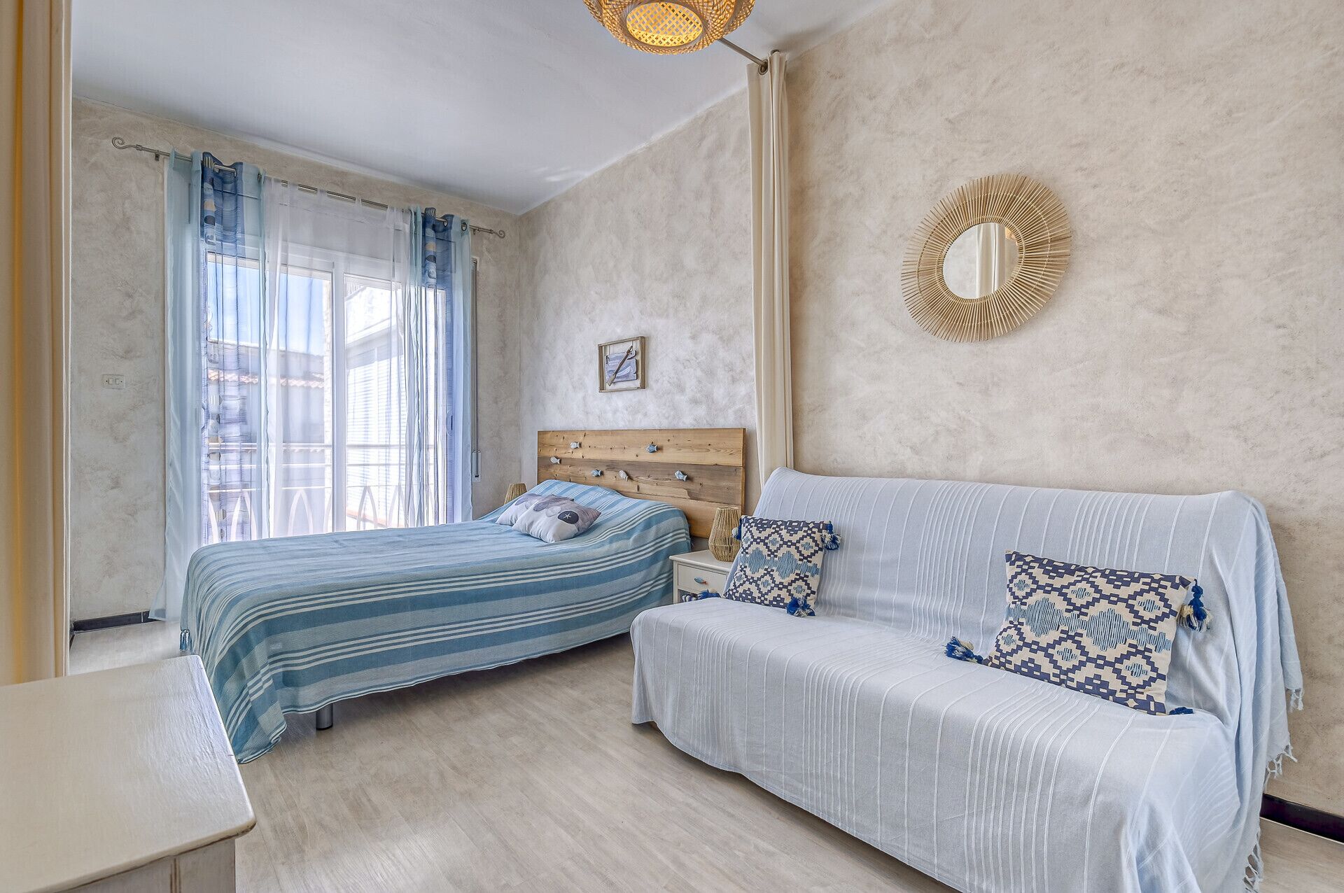 A bedroom with two beds, a sofa, a bedside table, a mirror, and a window with curtains.