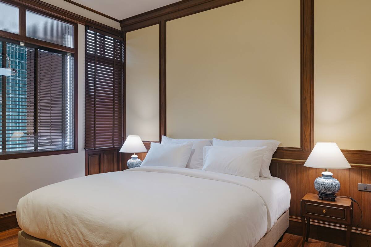 A neatly made bed with white linens, a bedside table with a lamp, and a window with blinds.