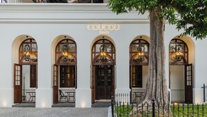 El exterior del Hotel Coucou, con tres ventanas en arco, cada una con una araña, y un gran árbol en primer plano.