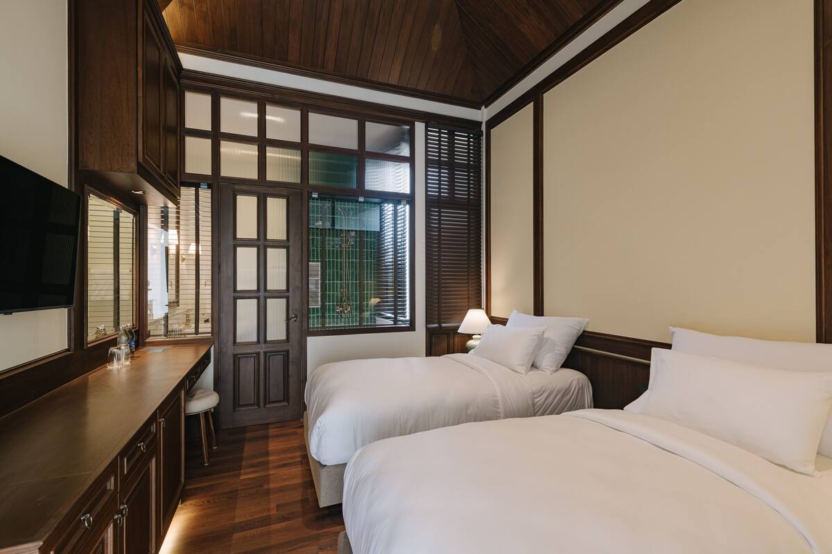 A hotel room with two beds, a wooden ceiling, a TV, and a window with blinds.