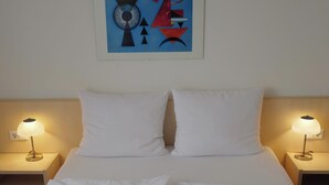 Een net opgemaakt bed met witte lakens, twee bedlampjes en een aan de muur hangend abstract schilderij.