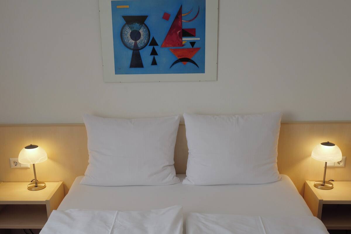 A neatly made bed with white linens, two bedside lamps, and a wall-mounted abstract painting.