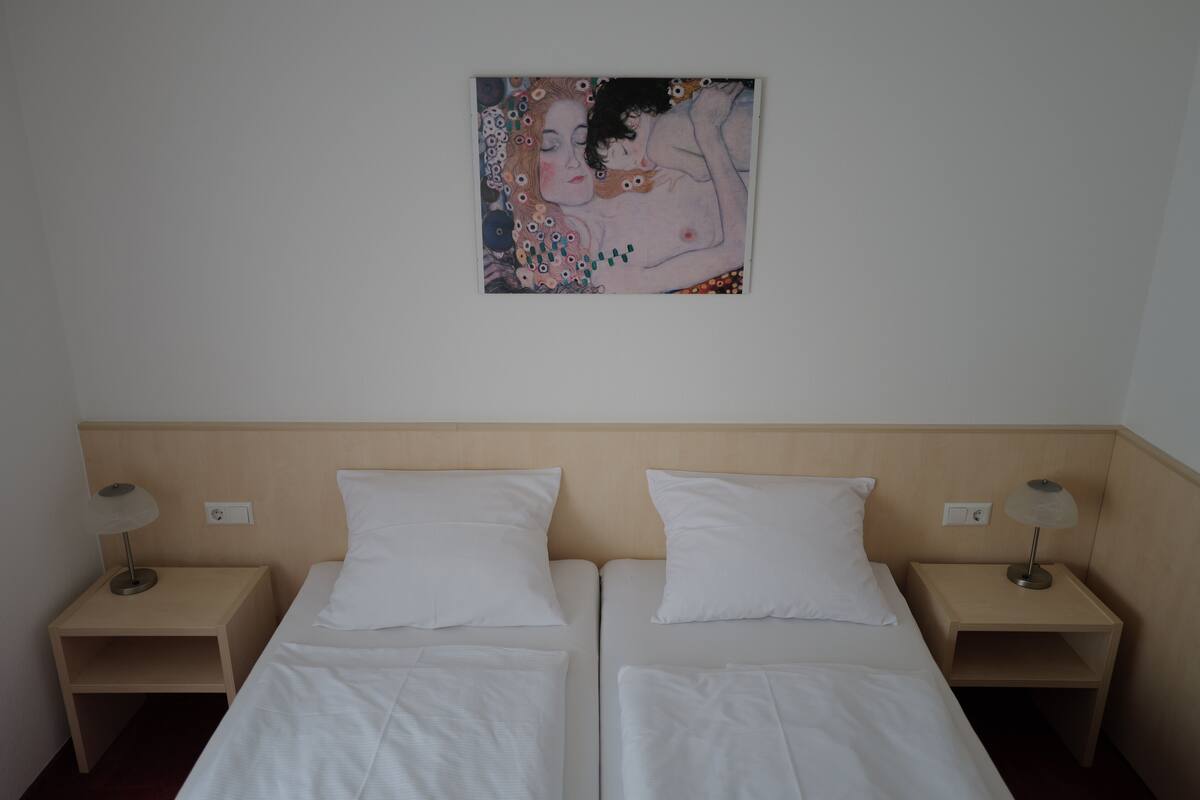 A hotel room with two beds, white linens, wooden headboard, bedside tables with lamps, and a painting on the wall.