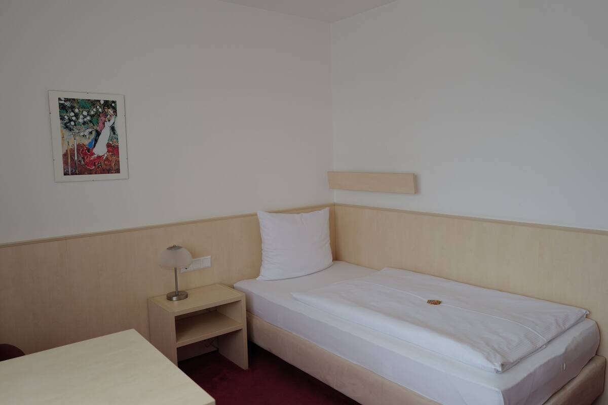 A small, single-bed room with a wooden headboard, a bedside table with a lamp, and a wall-mounted shelf.