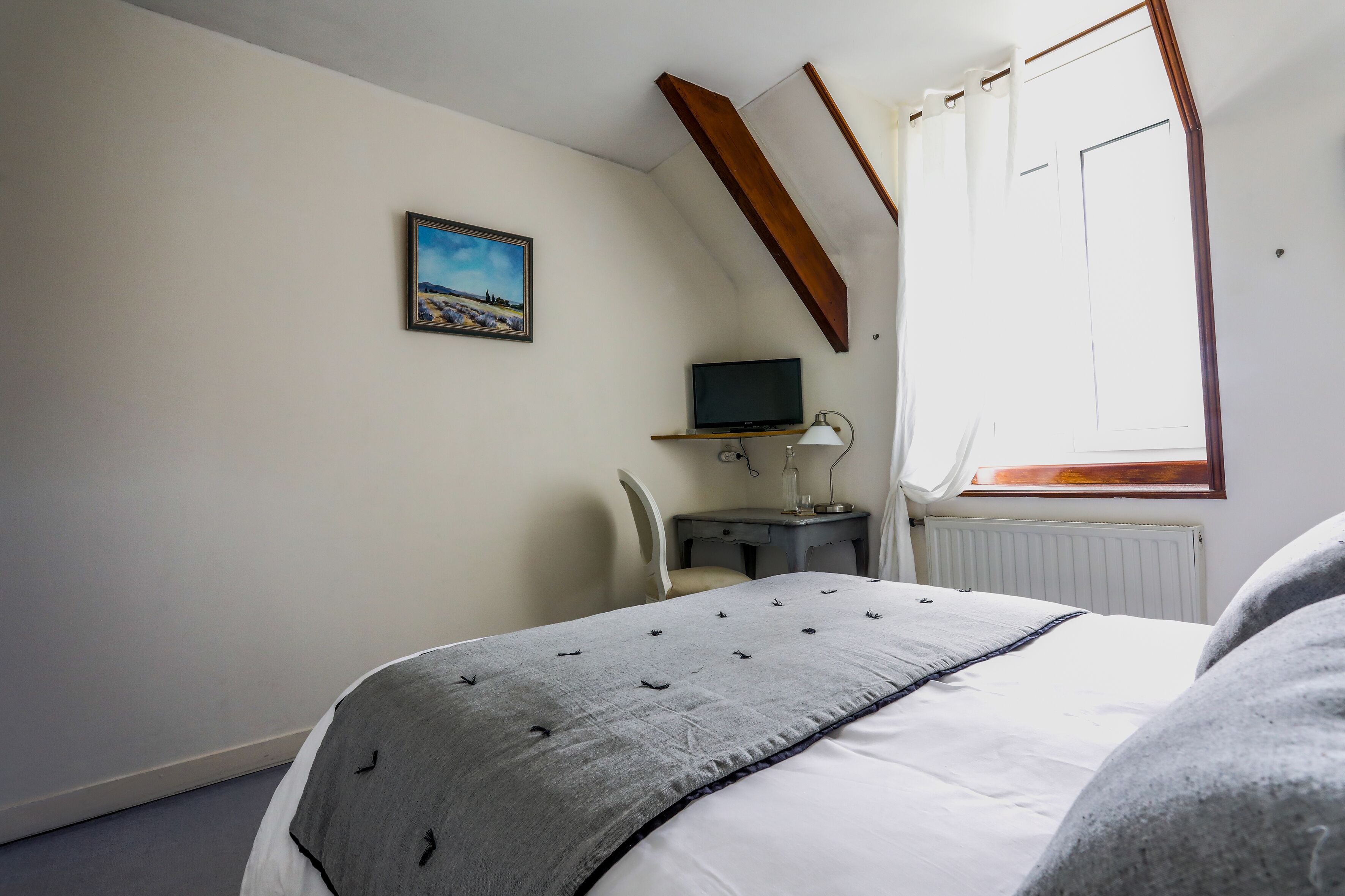 A bedroom with a bed, a desk with a computer, a chair, a window with curtains, and a painting on the wall.