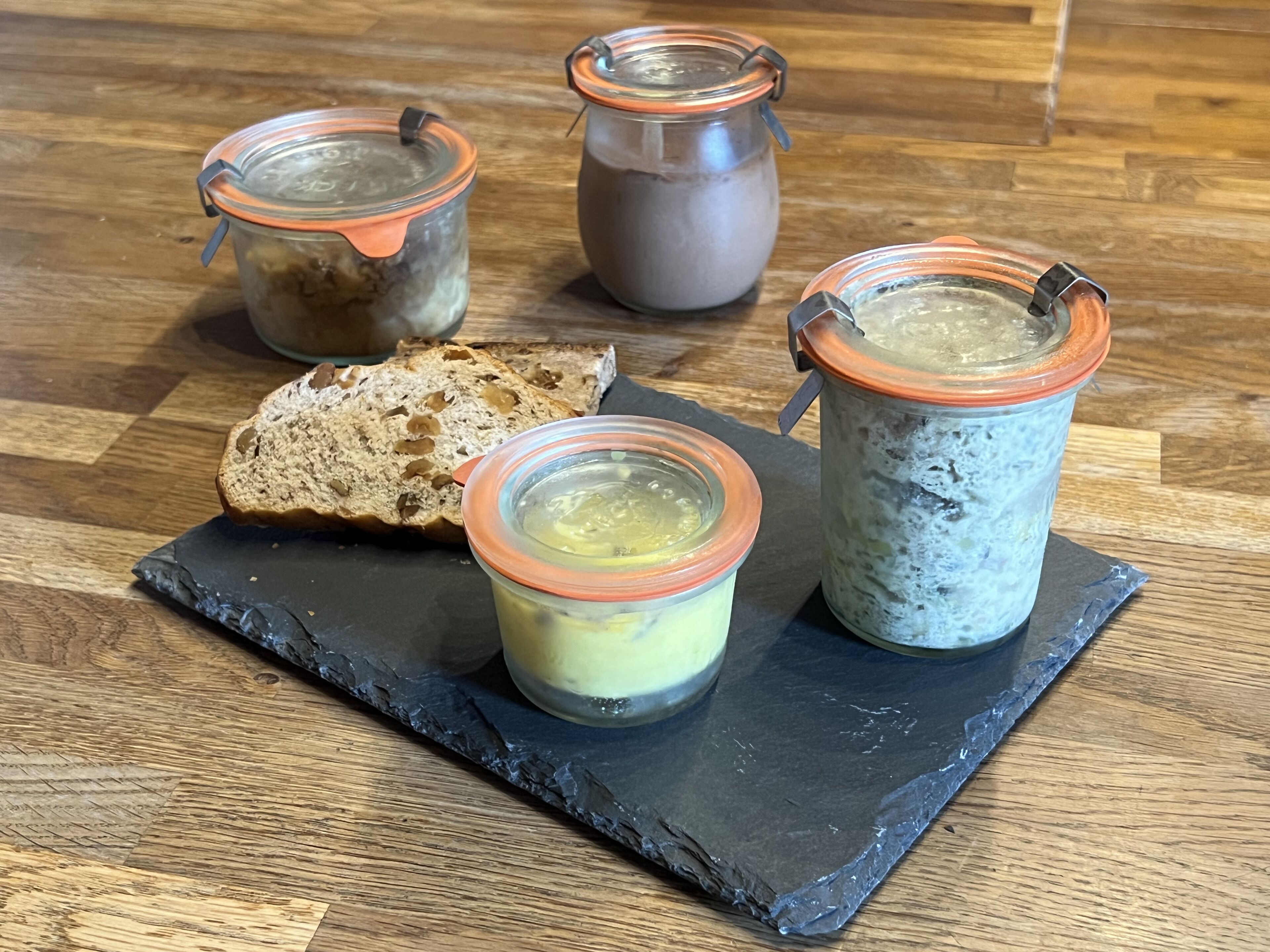A slate serving board with three jars of preserves and a slice of bread.