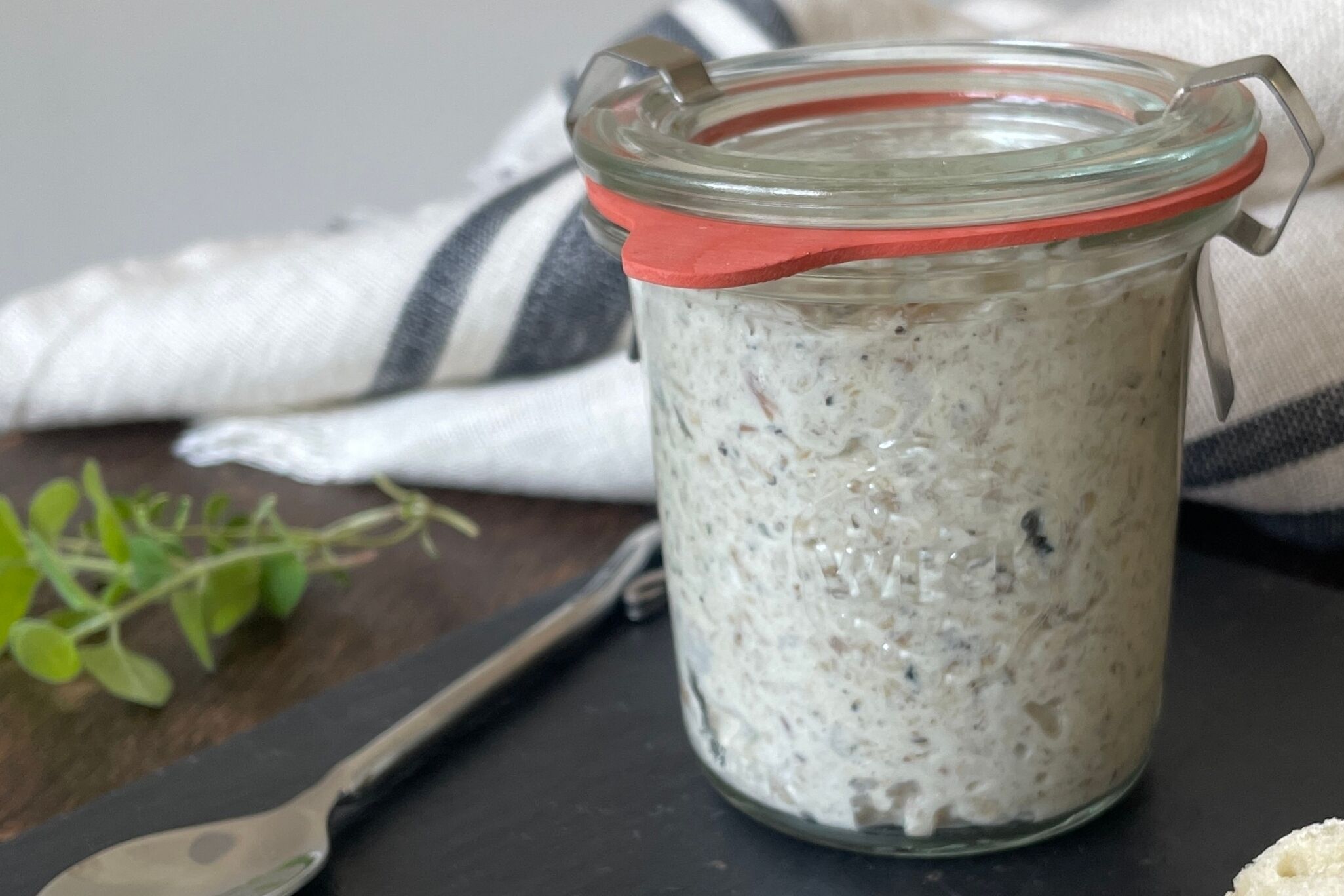 A jar of white creamy substance with specks, possibly a food item, on a dark surface with a spoon and a striped cloth nearby.