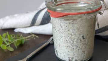 A jar of white creamy substance with specks, possibly a food item, on a dark surface with a spoon and a striped cloth nearby.