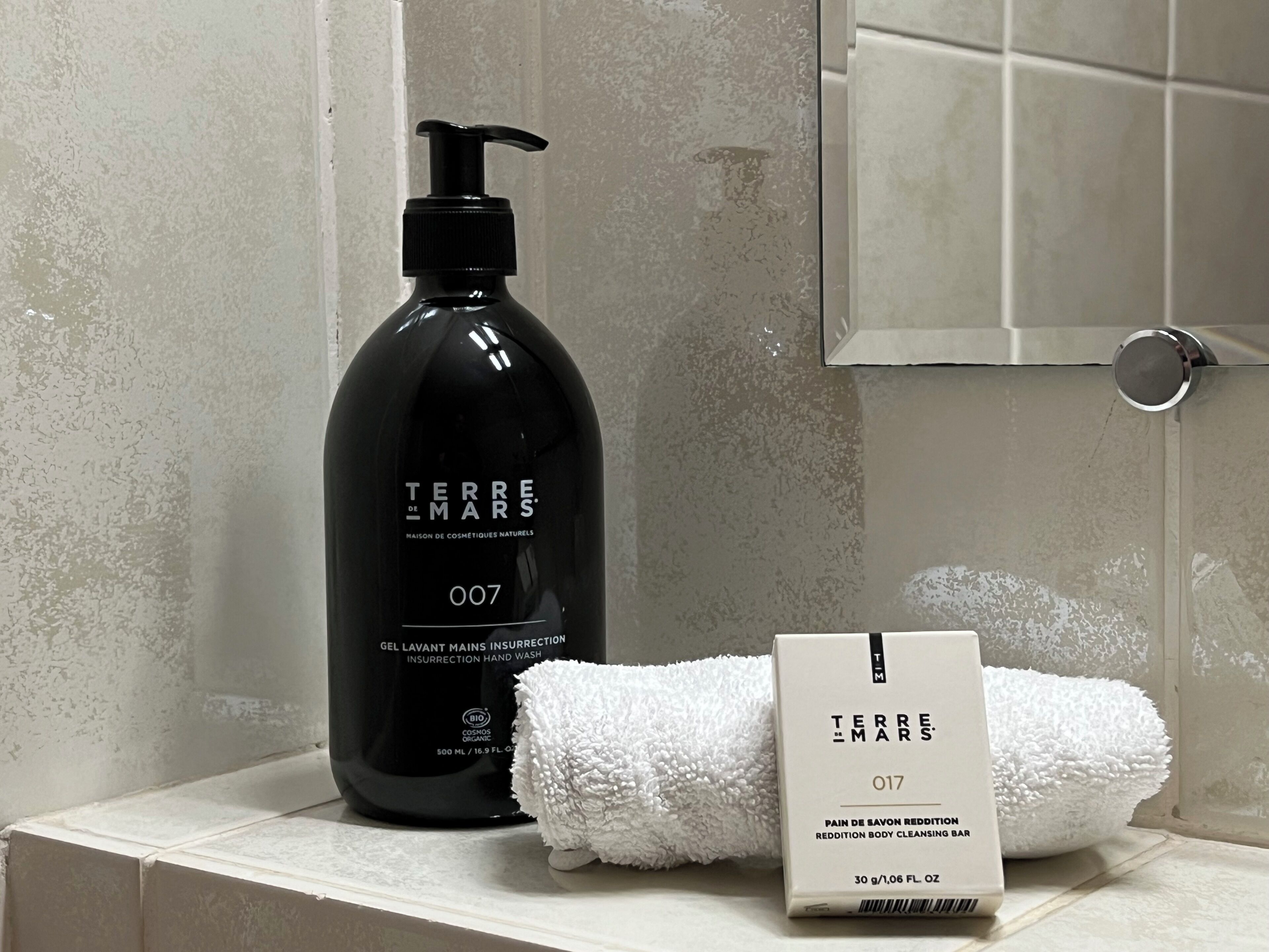 Baño con una botella negra etiquetada "Terre de Mars" y una toalla blanca sobre una repisa.