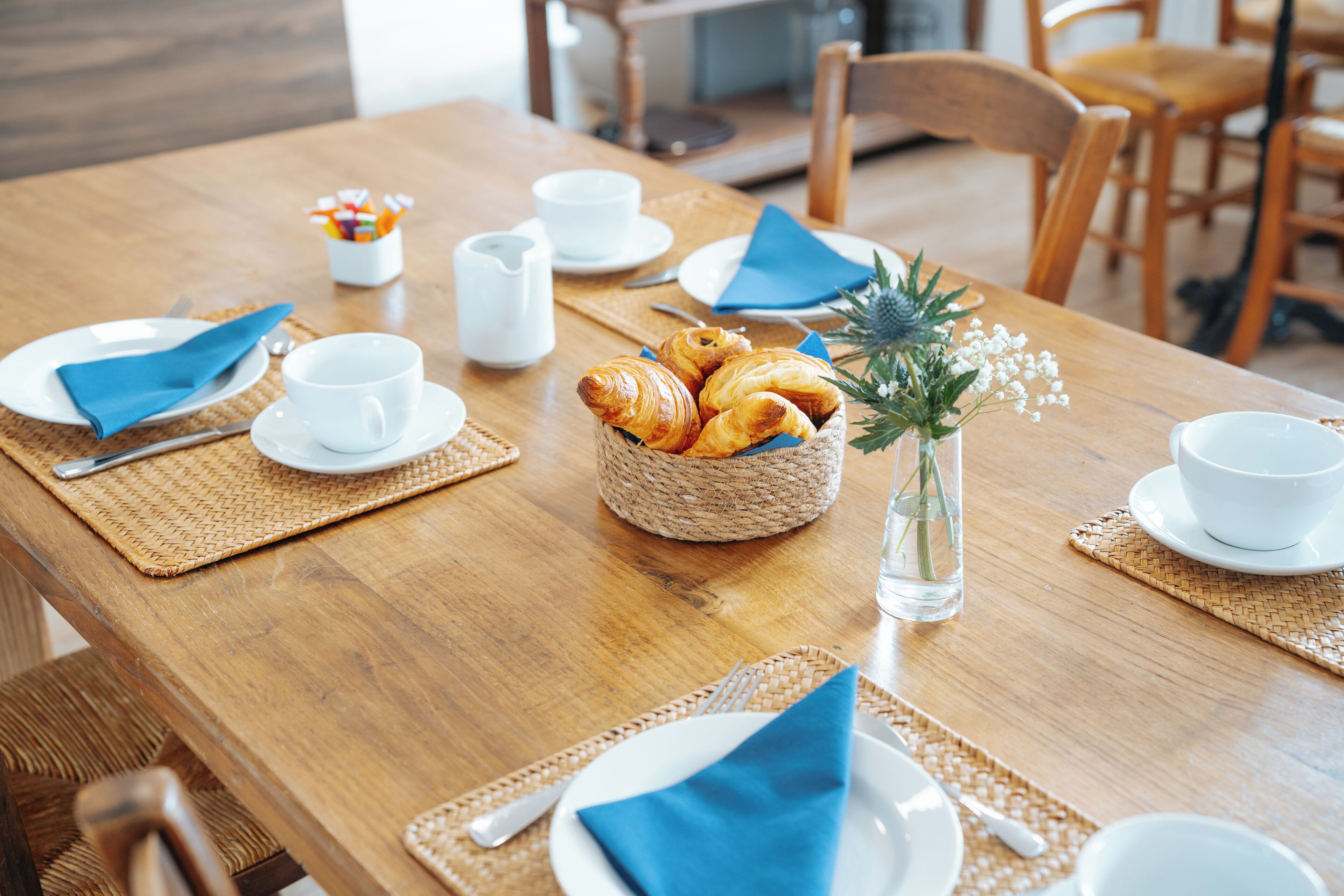 Een houten eettafel met borden, bekers en een mandje croissants.