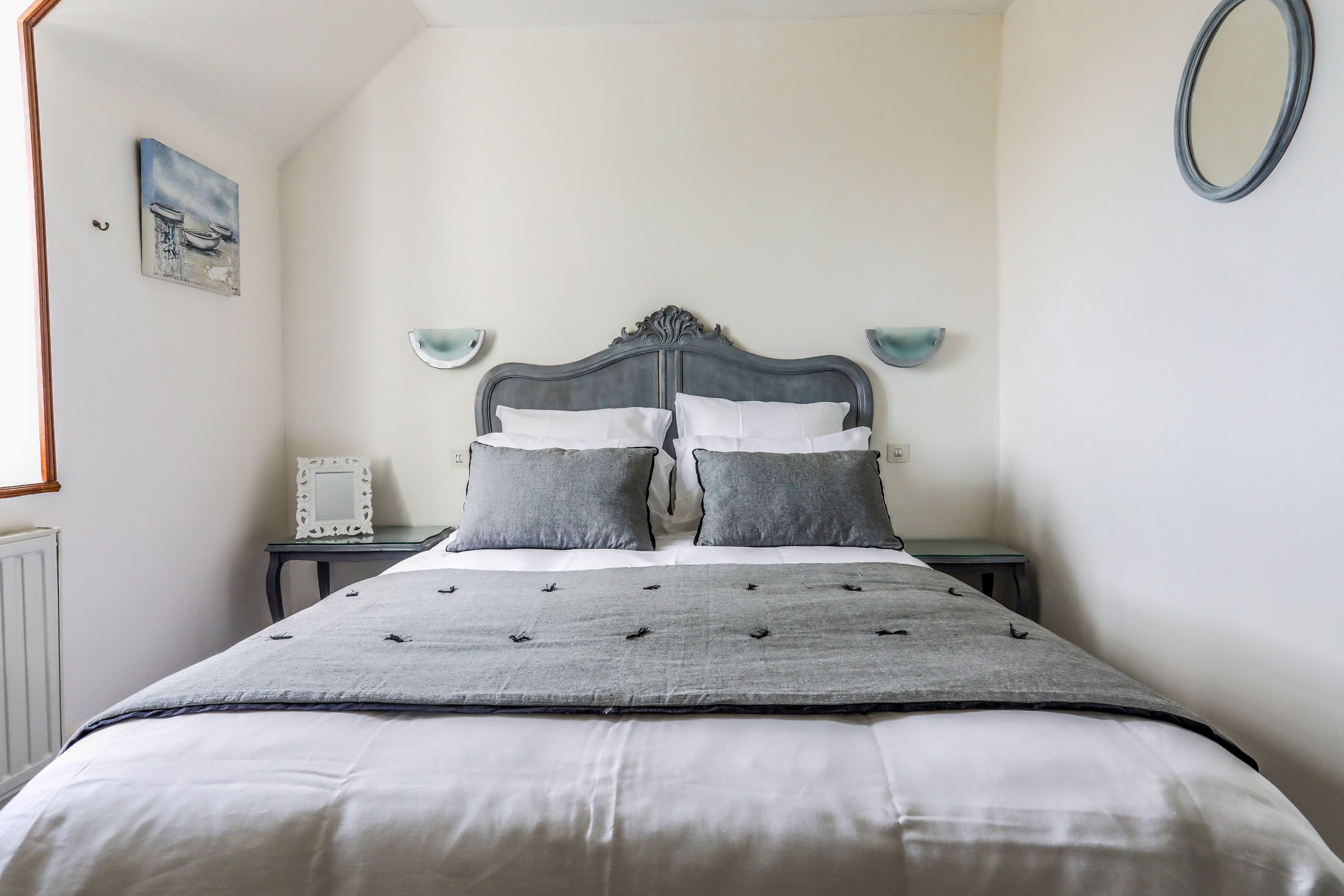 A bedroom with a bed, two bedside tables, a wall-mounted mirror, and a framed picture.