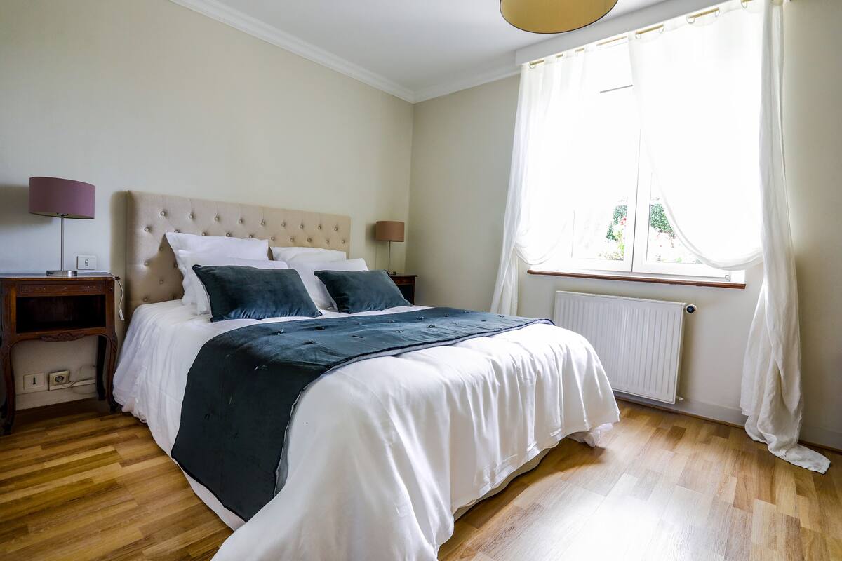 A bedroom with a large bed, a nightstand, a lamp, and a window with curtains.