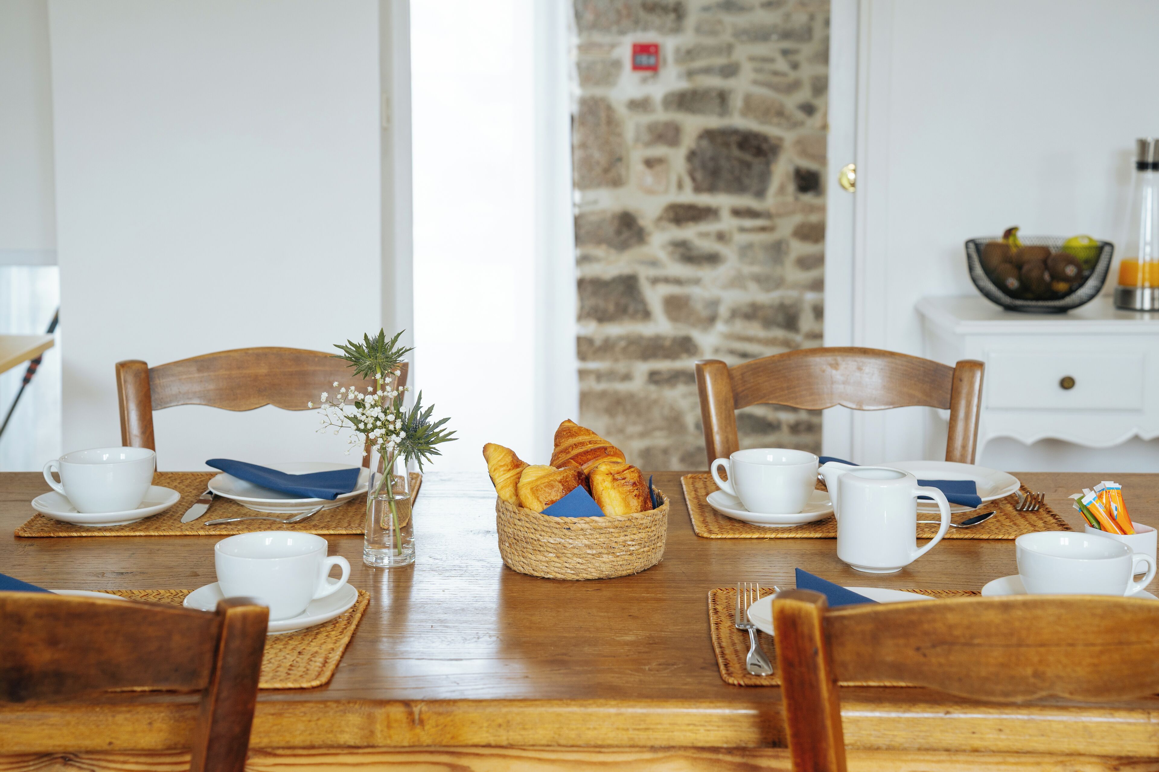 Une table de salle à manger en bois, avec des tasses et des soucoupes blanches, et un panier de pain.