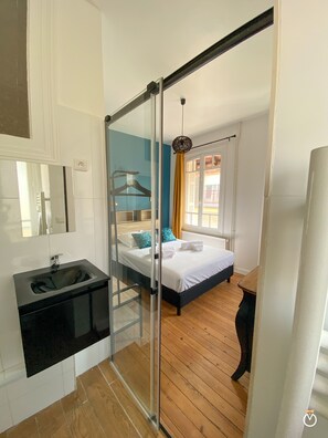 A bedroom with a bed, a bedside table, a window with curtains, and a bathroom with a sink and mirror. - La Rose des Sables (Trouville-sur-Mer)