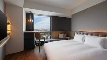 A modern hotel room with a large bed, a desk with a chair, a window with a city view, and a television on the left.