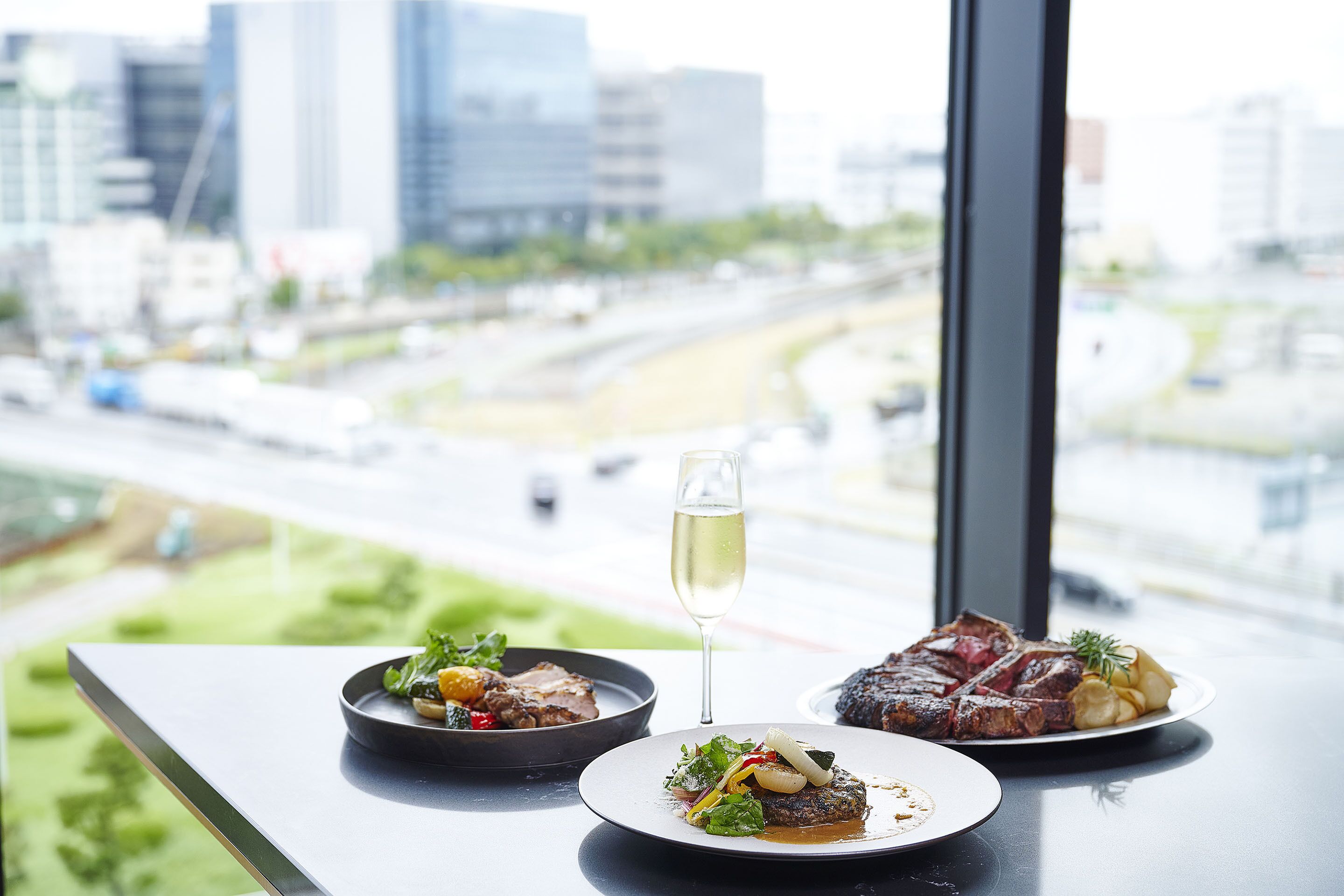 Une table avec deux assiettes de nourriture et un verre de vin, donnant sur un paysage urbain avec des immeubles et une autoroute.