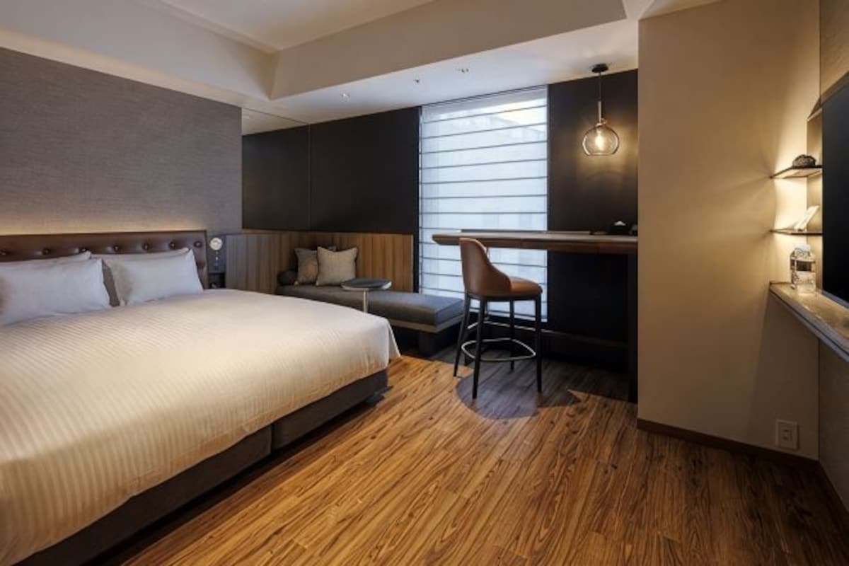 A hotel room with a large bed, a desk, a chair, and a window with blinds.