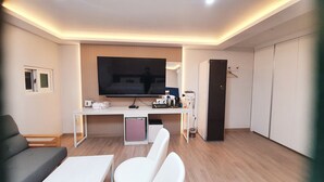 A modern hotel room with a flat-screen TV mounted on the wall, a white desk with a coffee maker, a mini-fridge, and a compact kitchenette. There is a sofa and two chairs in the seating area. - SWEET HOTEL (Changnyeong)