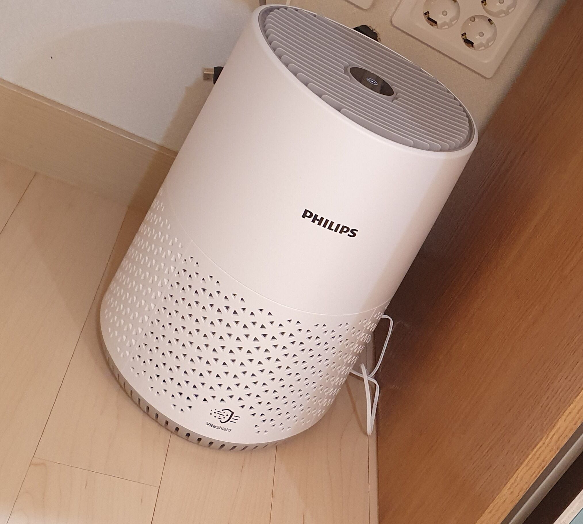 A white Philips smart speaker with a perforated base, placed on a wooden floor near a wall socket.