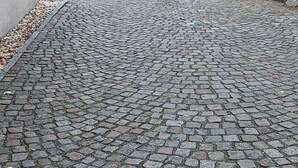 Ein Kopfsteinpflasterweg führt zu einem Gebäude mit einer Steinmauer und einem Hecken.