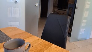 Ein modernes Wohnzimmer mit einem Holztisch, einer grauen Tasse und einem schwarzen Messer. Eine Schiebetür führt in eine Küche mit Backsteinwand und Fernseher.