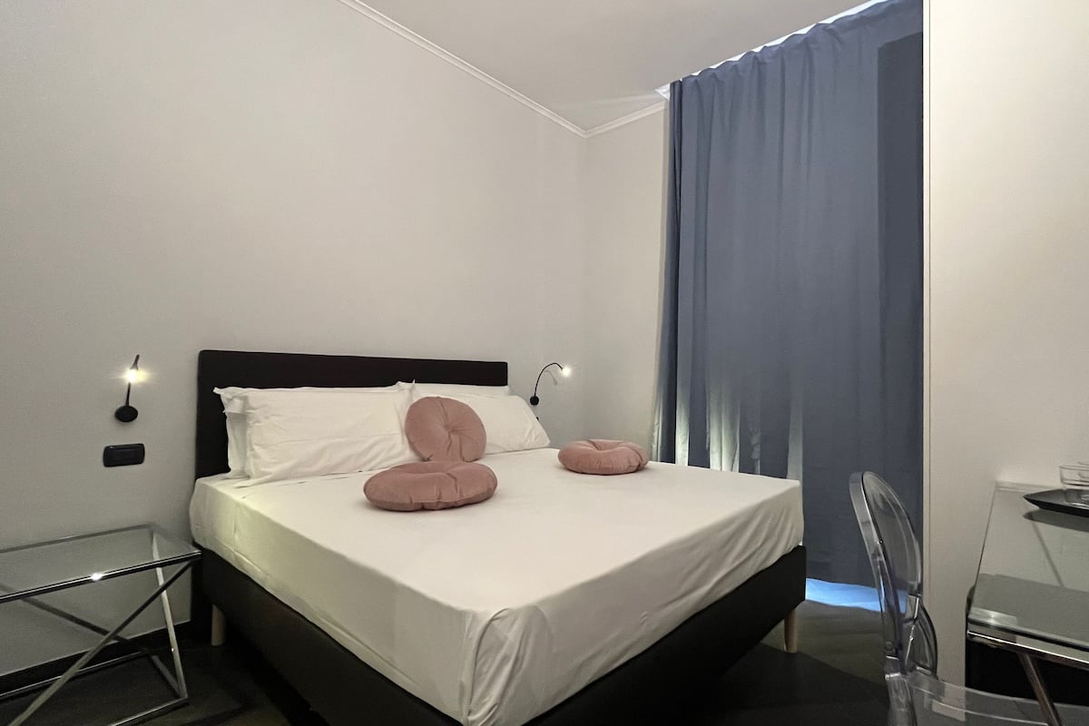 A modern hotel room with a bed, bedside tables, a desk, and a large window with curtains.