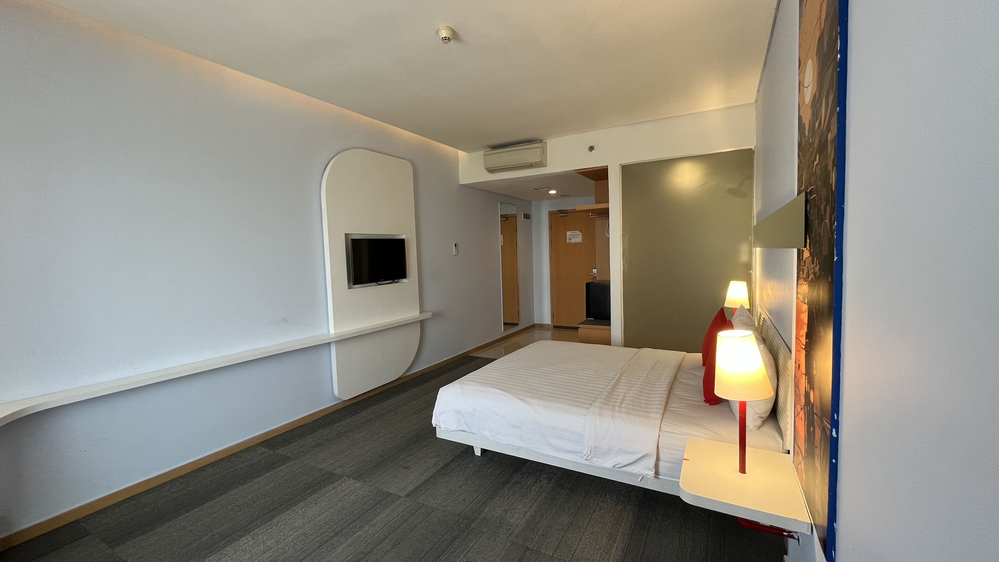 Een hotelkamer met een bed, een aan de muur bevestigde televisie, een bedlamp en een deur naar een andere kamer.