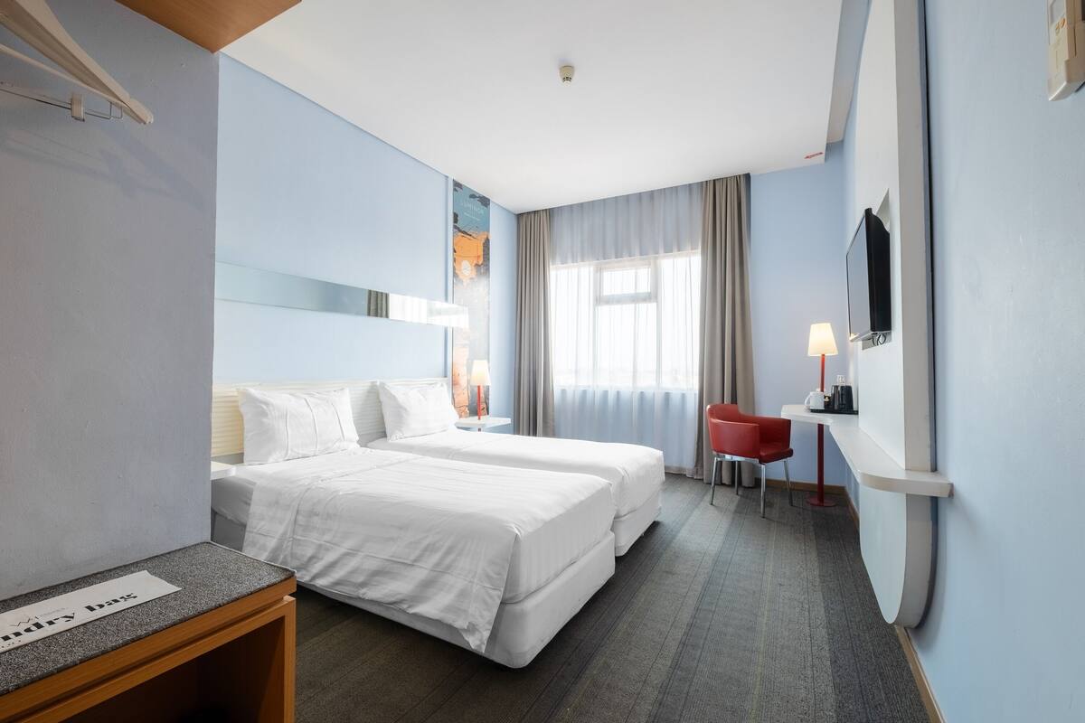 Une chambre d’hôtel avec deux lits, un bureau, une chaise, une télévision et une fenêtre avec des rideaux.