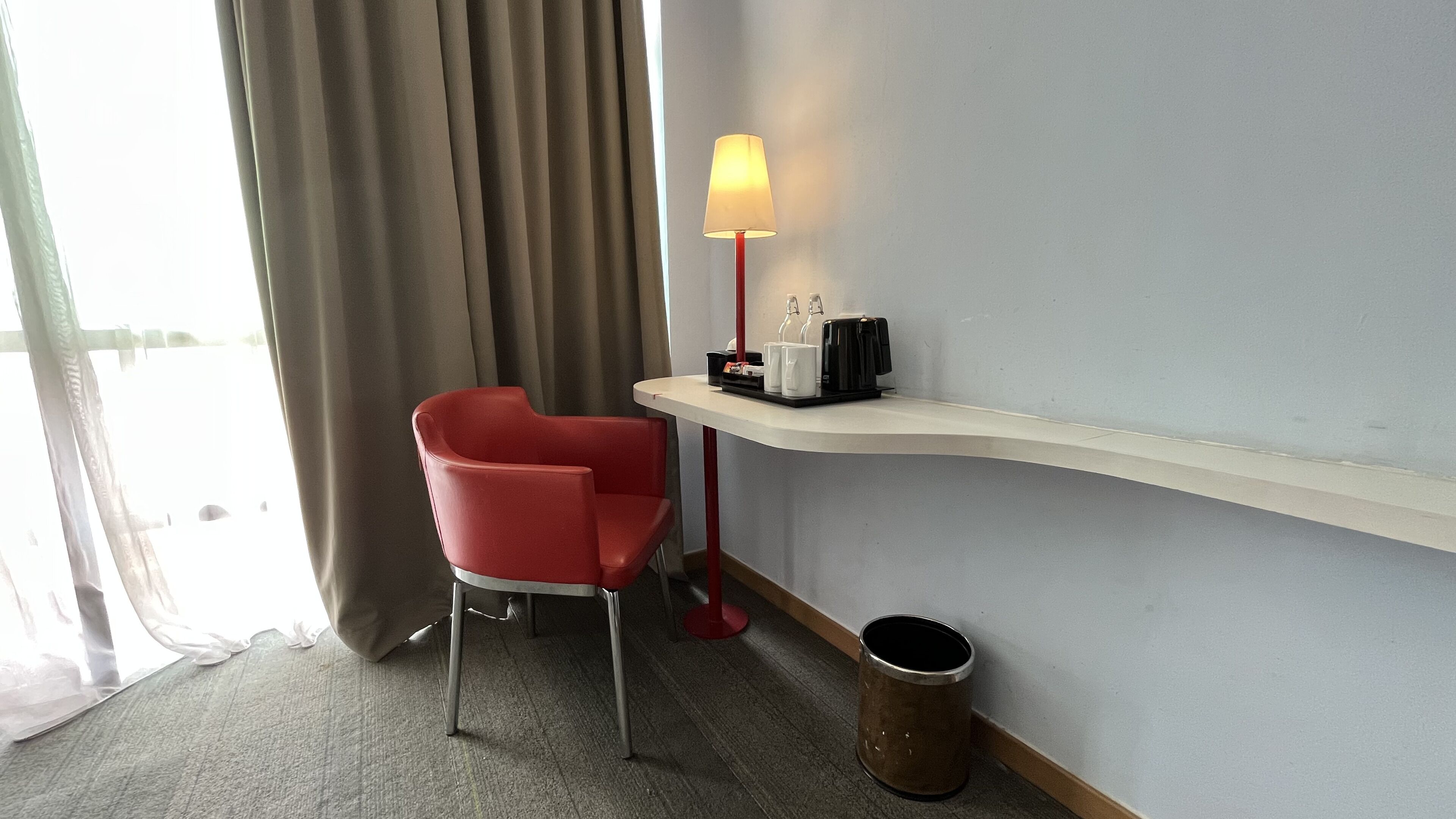 Une chambre d’hôtel avec un fauteuil rouge, une petite table avec une lampe, une poubelle et une fenêtre avec des rideaux.