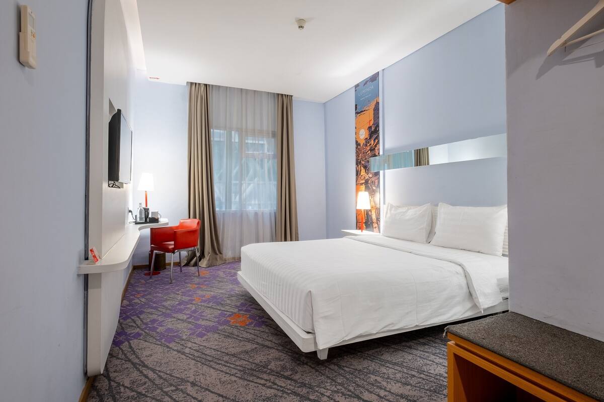 Une chambre d’hôtel avec un lit, un bureau, une chaise, une télévision et une fenêtre avec des rideaux.