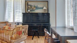 A room with a black piano, a painting of a coastal scene, a wicker crib with toys, and a wooden chair.