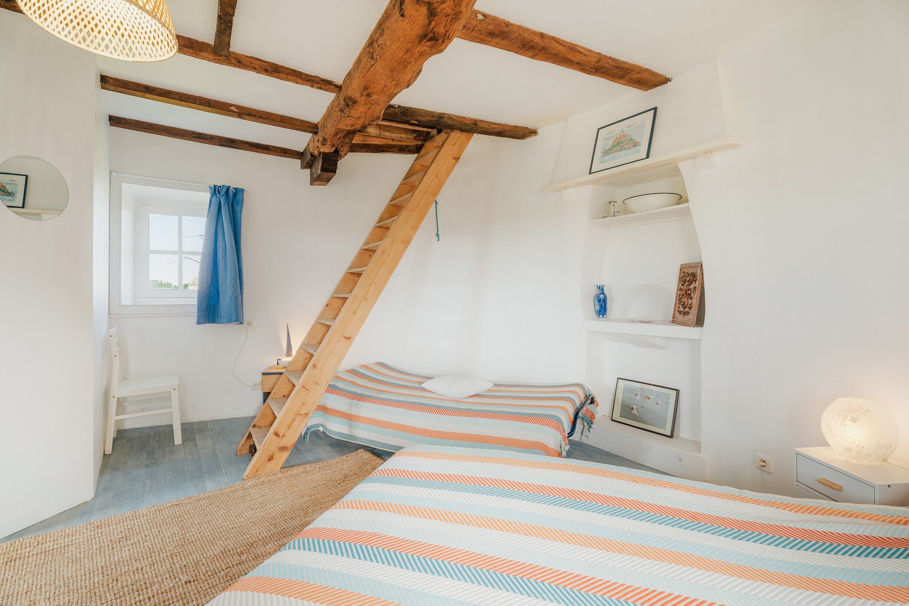 A bedroom with a sloped ceiling, wooden beams, a bunk bed, a desk, and a window with blue curtains.