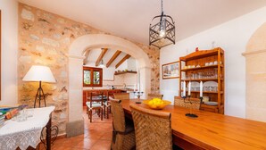 Un comedor con mesa de madera, sillas de mimbre, mantel blanco, lámpara y pared de piedra.