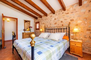 A bedroom with a bed, wooden nightstand, stone wall, and exposed wooden beams.