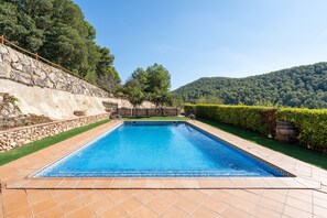 A rectangular swimming pool with a stone wall on one side and a wooden fence on the other, surrounded by greenery and hills in the background.