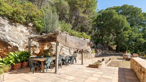 Un patio exterior con suelo de piedra, una pérgola de madera y una mesa con sillas.