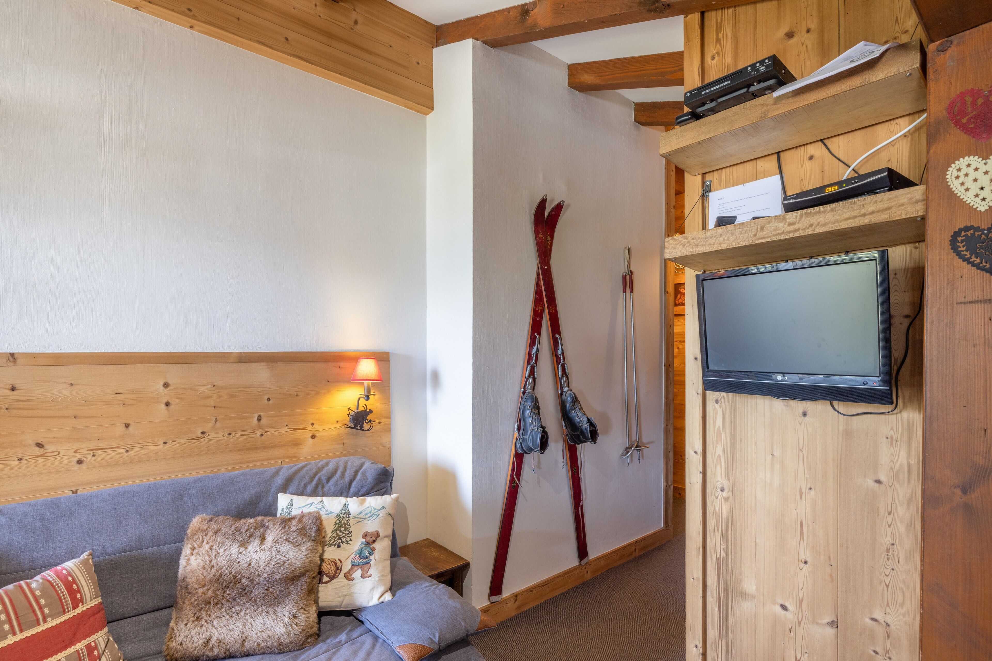 Een knus vertrek met een houten plafond, een grijze bank met decoratieve kussens, een bevestigde televisie en ski's die tegen de muur staan.