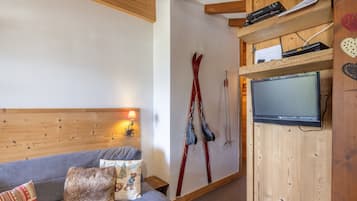 Een knus vertrek met een houten plafond, een grijze bank met decoratieve kussens, een bevestigde televisie en ski's die tegen de muur staan.