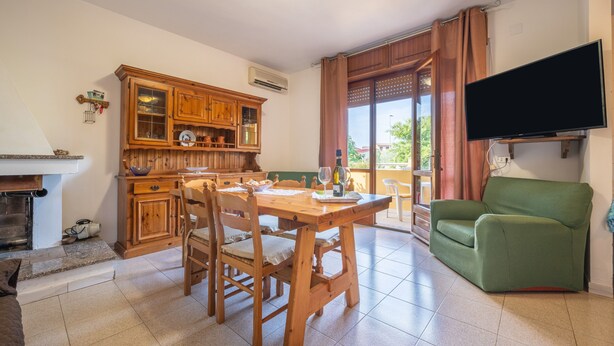 Een zithoek met een houten tafel en stoelen, een groene bank, een flatscreen-tv aan de muur, en een keuken met houten kastjes en een raam.