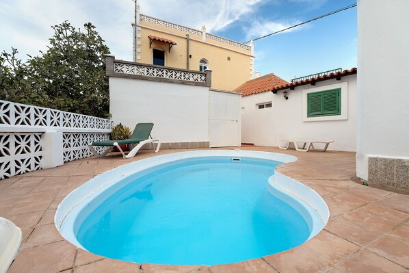 Une piscine avec une terrasse blanche, une clôture blanche et, au fond, un bâtiment doté d’un toit recouvert de tuiles.