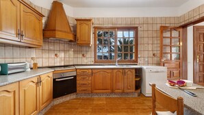 A kitchen with wooden cabinets, a built-in oven, a dishwasher, and a window with a view of palm trees. - Vacation Home 'Biofinca Las Dulces Villa Alma' with Sea View, Pool & Wi-Fi (Guia de Isora)