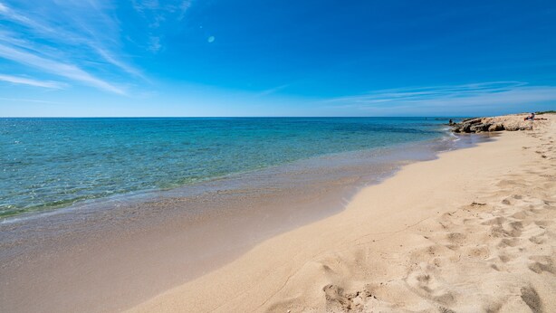 Une plage avec une eau d’un bleu limpide, un rivage sablonneux et des affleurements rocheux.
